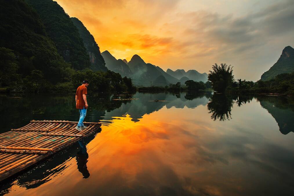 A man stands on a raft on a river with a yellow sunset and mountains in the background.