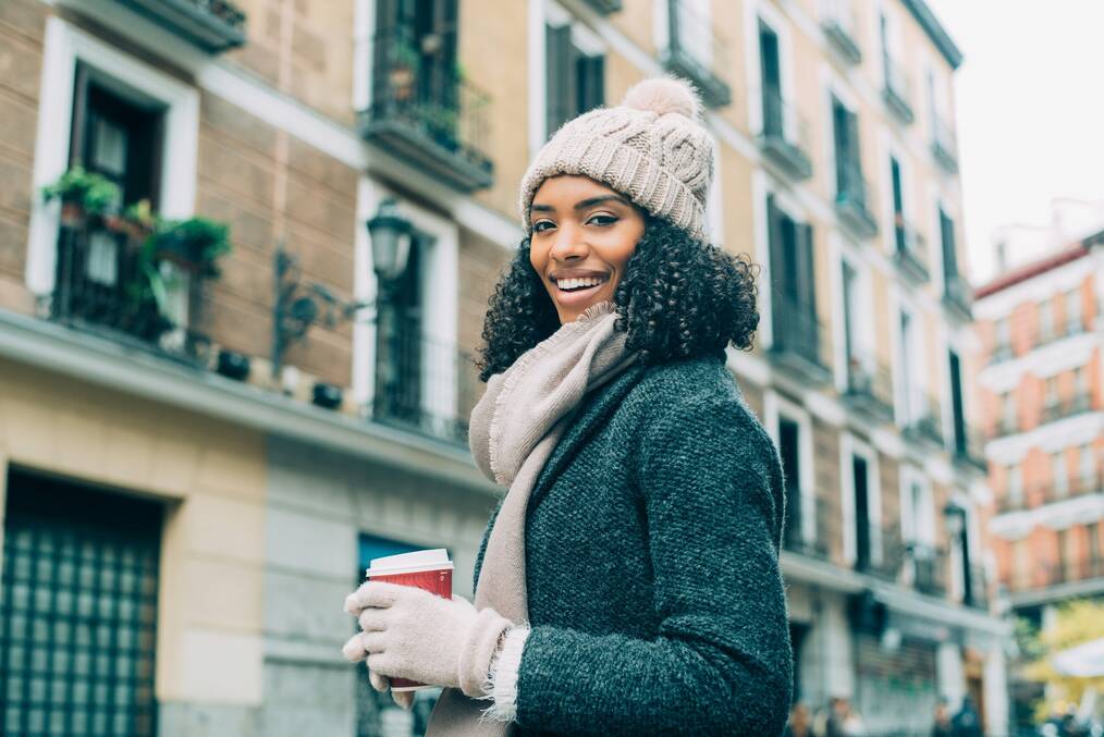A woman in a coat and hat poses in front of buildings in Madrid.