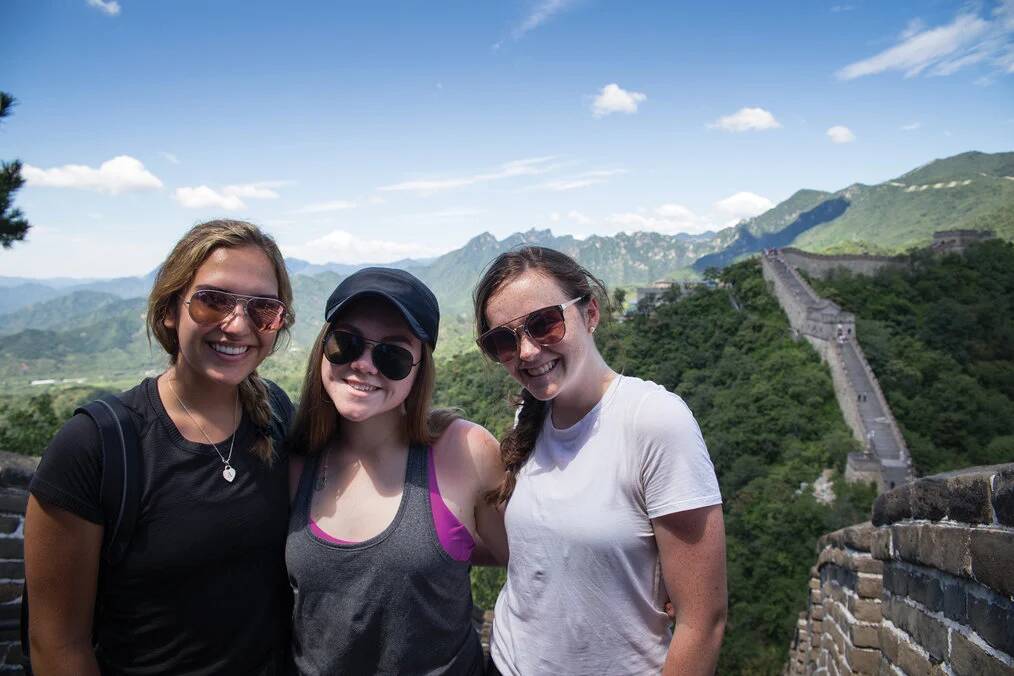 Three high school girls smile with the Great Wall of China in the background.