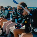 Students sitin the sun in a row on a boat with snorkel gear on, preparing to get into the turquoise waters under clear blue skies. 