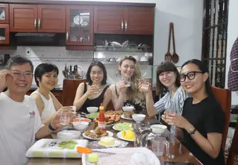 A group of six people sitting around a dining table, sharing a meal in a cozy kitchen setting. They are smiling and raising their glasses for a toast. The table is filled with a variety of dishes, including vegetables, shrimp, and chicken, as well as condiments and utensils. The kitchen in the background has wooden cabinets and shelves with dishes, creating a warm and inviting atmosphere. The group appears to be enjoying a pleasant and convivial moment together.