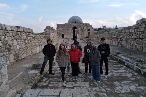 A group of 5 students stand in a row on an ancient paved road with ruins in the background.