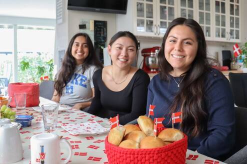 Three girls are sitting at a table and smiling. In front of them is a basket of baked good and Danish flags.