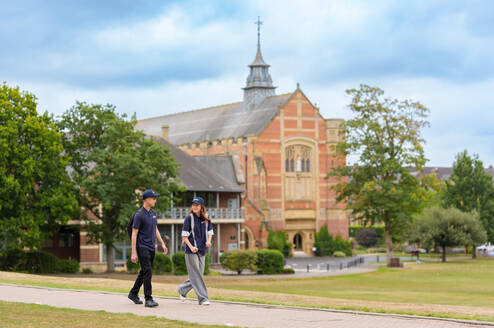 Two students walking on a paved path in front of a large historic building with a tall spire, surrounded by trees and manicured lawns.