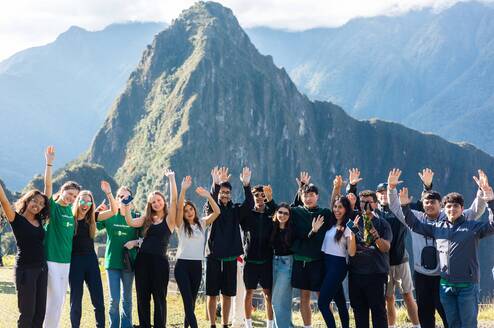 A group of teenagers standing on a stone terrace at Machu Picchu, waving their hands in the air with the ancient Incan ruins and Huayna Picchu mountain in the background.
