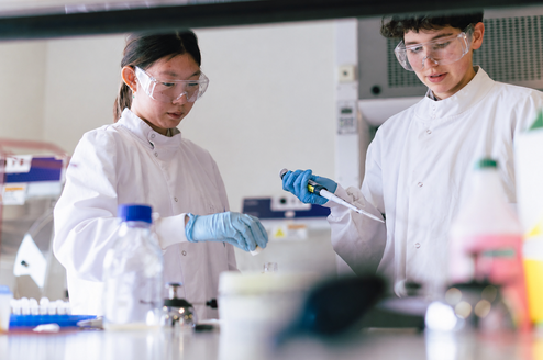 Two teenage students wearing lab coats, gloves, and safety goggles carry out a hands-on biomedical science experiment using a pipette in a modern laboratory setting.