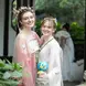 Two women in traditional attire, likely inspired by ancient East Asian fashion, are smiling and posing together in an outdoor setting with lush greenery. One is wearing a pink dress with elaborate hair accessories, and the other is in a white and green dress, holding a small blue and yellow plush toy. They stand close to a stone structure. The scene is vibrant and captures the beauty of cultural attire and natural surroundings.