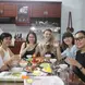 A group of six people sitting around a dining table, sharing a meal in a cozy kitchen setting. They are smiling and raising their glasses for a toast. The table is filled with a variety of dishes, including vegetables, shrimp, and chicken, as well as condiments and utensils. The kitchen in the background has wooden cabinets and shelves with dishes, creating a warm and inviting atmosphere. The group appears to be enjoying a pleasant and convivial moment together.