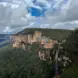 "A scenic view of a lush, green forested mountain range with sheer cliffs. The cliffs have a mix of greenery and exposed orange and brown rock faces. A waterfall cascades down the side of the cliff, blending into the dense forest below. The sky above is partly cloudy, with patches of blue sky peeking through."