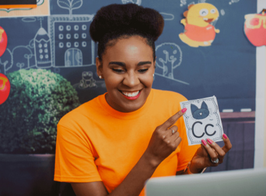 a woman in an orange shirt is smiling at a computer and pointing to a flashcard, which shows a picture of a cat and the letter C