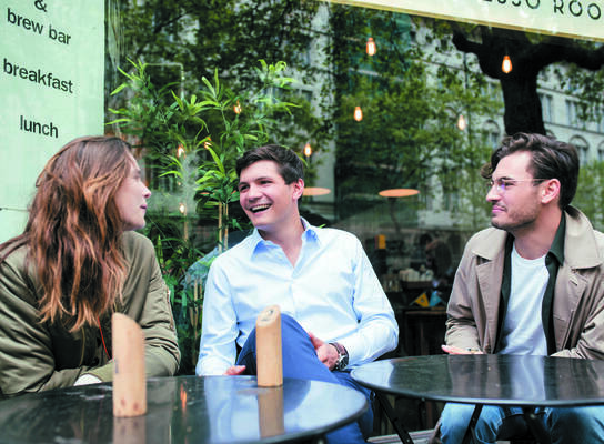 One female and two male postgraduate students sit at a table outside a cafe