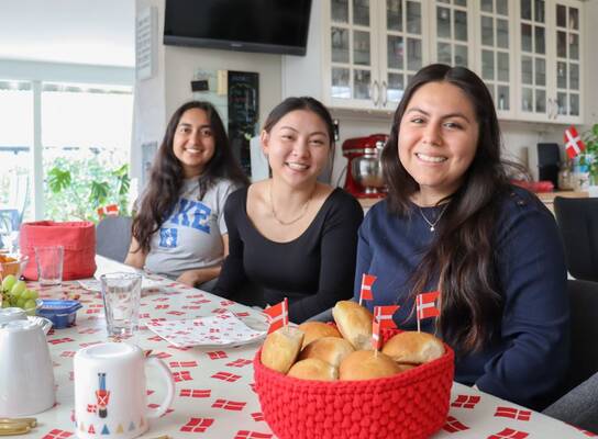 Three girls are sitting at a table and smiling. In front of them is a basket of baked good and Danish flags.