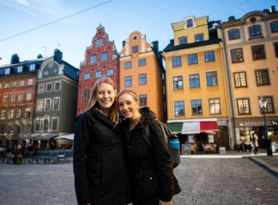 Two female students are standing in an old town square with colorful historical buildings behind them.