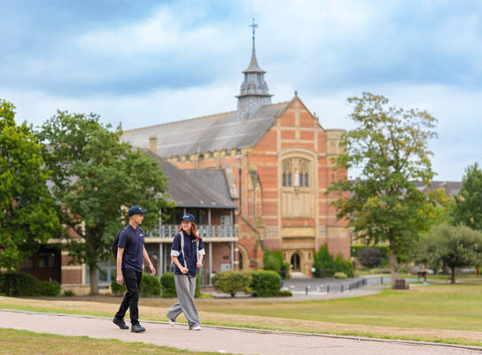 Two students walking on a paved path in front of a large historic building with a tall spire, surrounded by trees and manicured lawns.