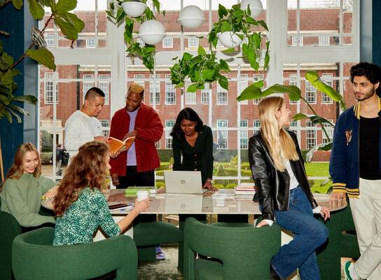 A group of students standing around a long table in front of a large white-pane window. The students are looking through text books, working on laptops, drinking coffee and chatting and laughing with one another. 