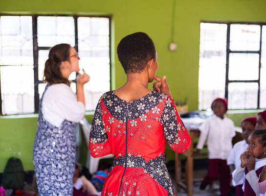 A volunteer teacher and a local student stand before a classroom of young children in Tanzania. They are pointing to their mouths to demonstrate pronunciation against a backdrop of bright green walls.
