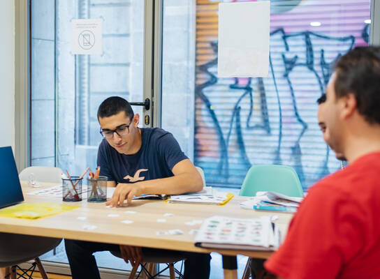 Students sitting around a table in a classroom, studying Spanish together with books and learning materials in a bright, modern setting.