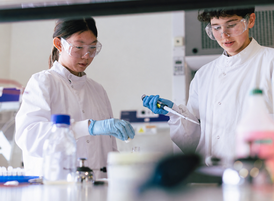 Two teenage students wearing lab coats, gloves, and safety goggles carry out a hands-on biomedical science experiment using a pipette in a modern laboratory setting.