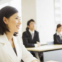 smiling Asian woman wearing a white suit sitting at a desk with two other people wearing suits in the background