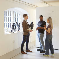 A tutor talks to two students standing by a sculpture