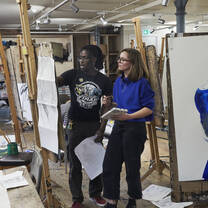 A tutor speaks to a young student in front of an easle