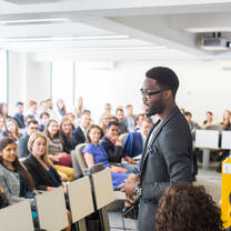 A male student stands before a classroom of postgraduate students