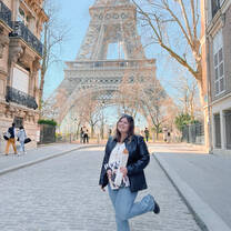 A young woman in a black jacket stands in front of the Eiffel Tower with a baguette and a magazine in hand