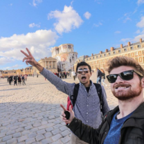 Two male students stand in front of a long historical building, both holding up peace signs at the camera.