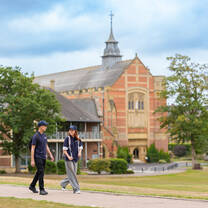 Two students walking on a paved path in front of a large historic building with a tall spire, surrounded by trees and manicured lawns.