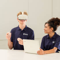 Two students seated at a table; one wearing a virtual reality headset and holding controllers, the other using a laptop. Both wear matching IMPACT polo shirts.