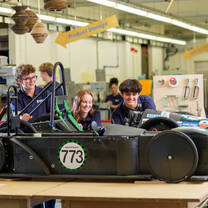 Three students in a workshop examining a black race car numbered 773, surrounded by tools and equipment with visible signage like 'Product Storage.'