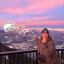 A girl in front of a mountain at sunset pointing looking shocked. There is a bear illustration added.