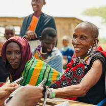 A smiling Maasai woman in traditional red clothing receives a medical checkup from a healthcare worker at an outdoor community outreach clinic in Tanzania