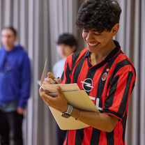 A boy in a football tshirt laughs as he looks down at his script. He is standing in a studio with two blurred people behind him.