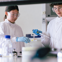 Two teenage students wearing lab coats, gloves, and safety goggles carry out a hands-on biomedical science experiment using a pipette in a modern laboratory setting.